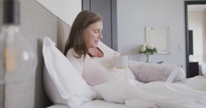 Woman drinking coffee while having a video chat on her laptop in bed at home