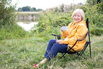 Happy middle aged blonde woman traveler in yellow hoodie holding iron mug cup with tea or coffee, sitting in chair at campsite near lake or river, smiling looking at camera, enjoying nature landscape