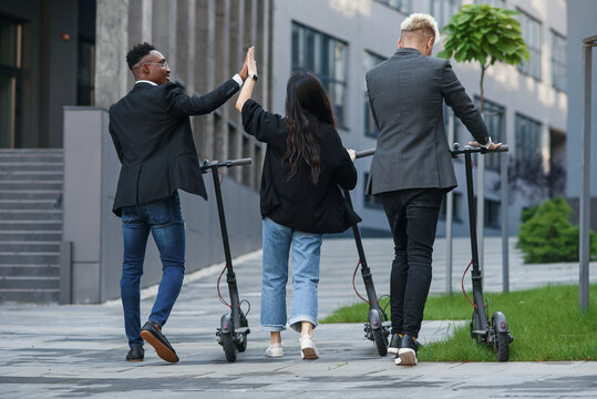 Confident multiracial office colleagues discussing business project go near office building with electric scooters