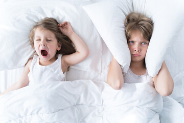 Little girls sleep in bed. One is snoring hard, sister is plugging her ears with pillow. Early...