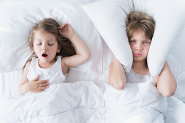 Little girls sleep in bed. One is snoring hard, sister is plugging her ears with pillow. Early...
