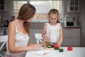 Happy family mother and child posing at home. Beautiful young mom and little daughter having fun and preparing vegetables for salad in a white kitchen in a Scandinavian style interior. Healthy food.