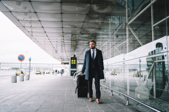 Caucasian businessman with luggage suitcase walking at airport exterior getting to departure terminal, formally dressed male traveller 30 years old with baggage ready for work trip by airplane