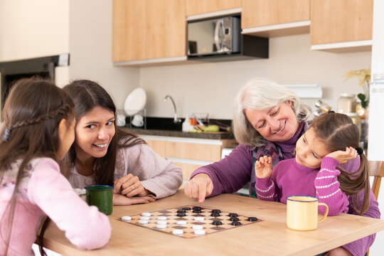 Joyful Retired grandmother with game board teaching granddaughter to play in kitchen home, indoors. Relationship, leisure, enjoyment, aging concept.