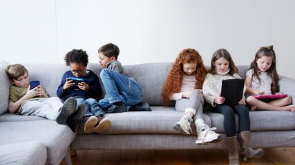 Group of children with friends sitting on sofa at home playing together on handheld computer game digital tablet and mobile phone - shot in slow motion