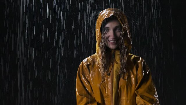 Attractive young woman in yellow raincoat stands on the street at night in the rain. Cute girl straightens her hood so as not to get wet and looks at the camera.