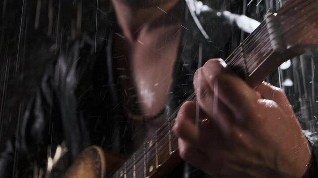 Close up hands sexy guitarist in black leather jacket plays acoustic guitar and sings song on black background in the rain outside at night.