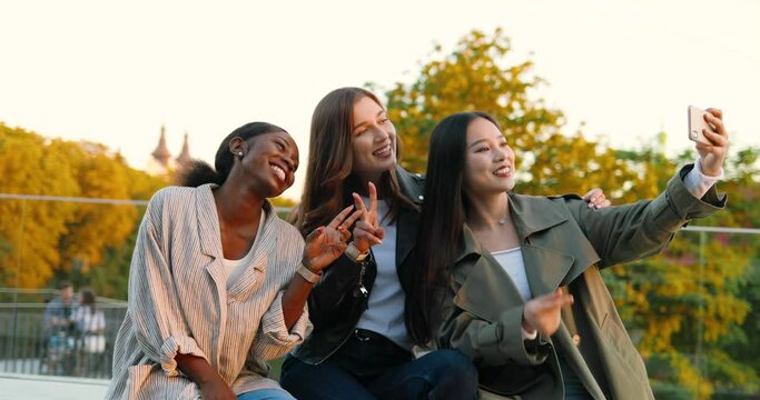 Cheerful Caucasian, Asian, African American beautiful females posing and smiling to smartphone camera. Mixed-races young women taking selfie photo with phone. Multi ethnic girls making photos outdoors