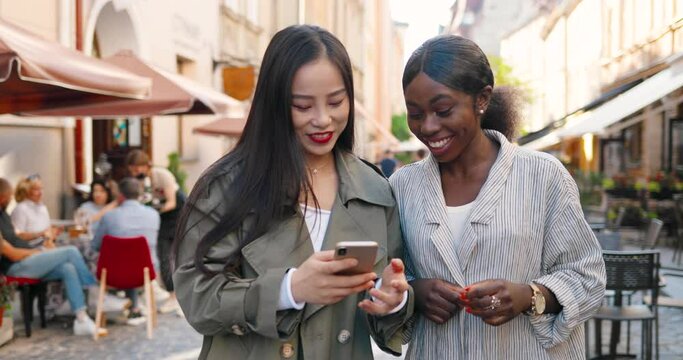 Mixed-races females friends walking the street, talking and watching video on phone. Multi ethnic young women chatting and using smartphone. Joyful Asian and African American girls gossiping. Gossips
