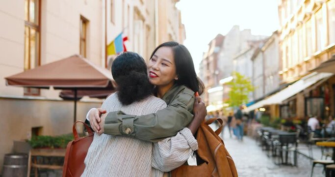 Young mixed-races beautiful joyful women meeting at street and hugging happily. Multi ethnic pretty females in hugs laughing and talking outdoor. African American and Asian girls best friends embrace.