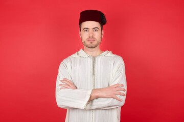 Obraz premium Portrait of serious young caucasian muslim man wearing djellaba and traditional hat over red background feels cool and confident entrepreneur crossing arms 