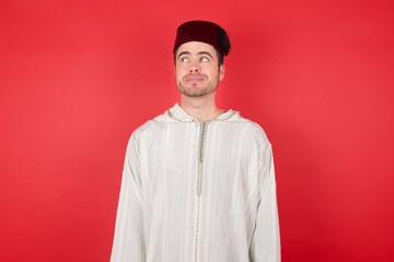 Obraz premium Photo of cheerful positive cute young caucasian muslim man wearing djellaba and traditional hat over red background, looking aside into empty space thoughtful