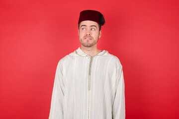 Photo of amazed young caucasian muslim man wearing djellaba and traditional hat over red background bitting lip and looking up to empty space, 