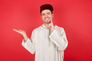 Funny young caucasian muslim man wearing djellaba and traditional hat over red background hold open palm new product great proposition 