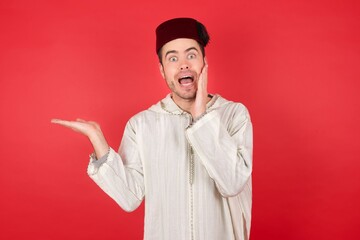Obraz premium young caucasian muslim man wearing djellaba and traditional hat over red background with an hairstyle advising discount prices hold open palm new product