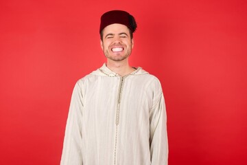 young caucasian muslim man wearing djellaba and traditional hat over red wall very happy and excited about new plans.