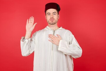 young caucasian muslim man wearing djellaba and traditional hat over red wall Swearing with hand on...