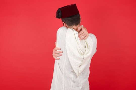 Young Caucasian Muslim Man Wearing Djellaba And Traditional Hat Over Red Wall Hugging Herself Happy And Positive From Backwards. Self Love And Self Care