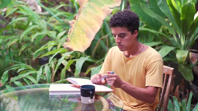 Latino digital nomad man writing in journal and looking up with an epiphany in lush tropical cafe on a sunny day