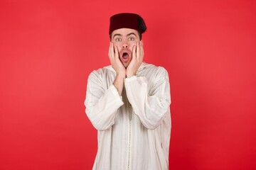 Stupefied Portrait of displeased upset young caucasian muslim man wearing djellaba and traditional hat, expresses excitement and thrill, keeps jaw dropped, hands on cheeks, has eyes popped out,
