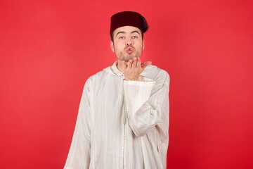 Portrait of displeased upset young caucasian muslim man wearing djellaba and traditional hat over red back looking at the camera blowing a kiss with hand on air being lovely and sexy. Love expression.