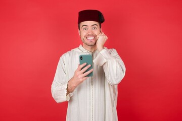 Joyous young caucasian muslim man wearing djellaba and traditional hat over red wall poses with mobile phone device, types text message on modern smartphone, watches funny video during free time, 