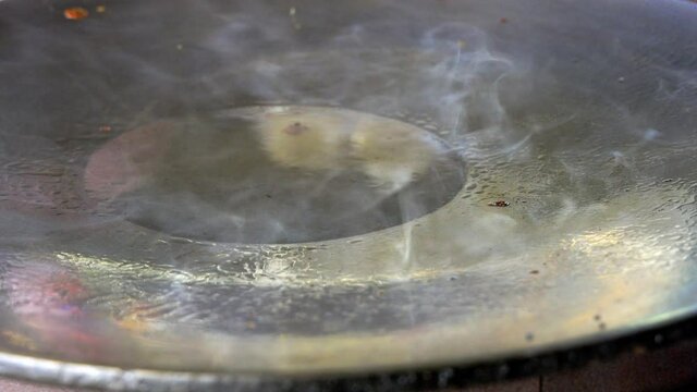 Street food vendor is heating oil in very hot pan with white smoke floating all over.