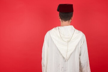 The back view of a young caucasian muslim man wearing djellaba and traditional hat over red background Studio Shoot.