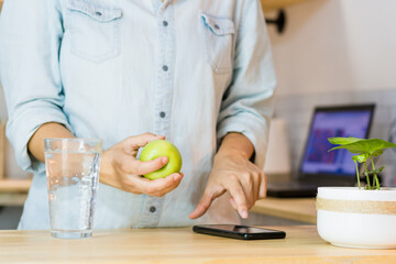 Woman drinking water while using her mobile phone, a fruit on the table.