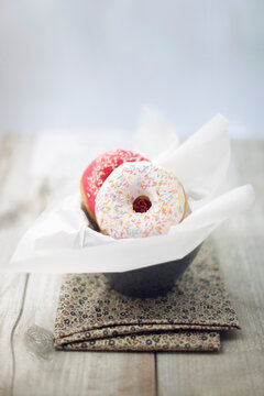 White and pink doughnuts with sugary on them in a cup on flowered cloth on wooden table
