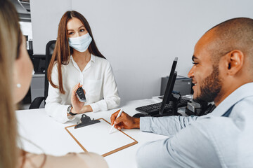 Attractive woman car seller wearing mask gives keys to car buyer