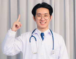 Portrait closeup of Happy Asian young doctor handsome man smiling in uniform and stethoscope neck strap pointing finger to side away and looking to the camera, healthcare medicine concept