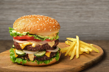 Grilled tasty beef burger with french fries on wooden rustic table against light wood wall background. Huge cheeseburger or hamburger with two bbq meat patty, lettuce, tomato, onion, pickles and sauce