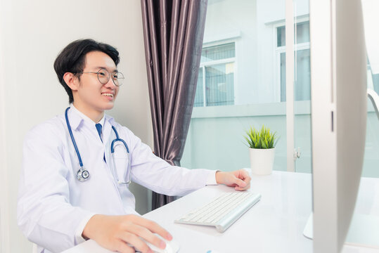 Asian Young Handsome Doctor Man Wearing A Doctor's Dress And Stethoscope Video Conference Call Or Facetime With Computer To Patient He Smiling Sitting On Desk At Hospital Office, Health Medical Care