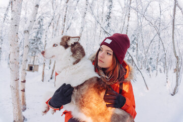 girl in a bright orange jacket sits and plays and hugs with a husky breed dog in the winter forest after snowfall during frost in the rays of the bright sunset sun