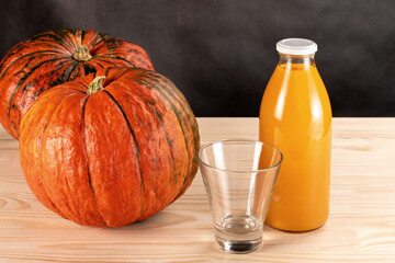 two pumpkins and a bottle of pumpkin juice on a wooden table. empty juice glass. dark grey background