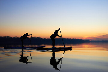 Fototapeta premium Silhouettes of two women paddle on stand up paddle boarding (SUP) on quiet winter or autumn Danube river at sunset. Colorful sunset over the river with silhouettes of people