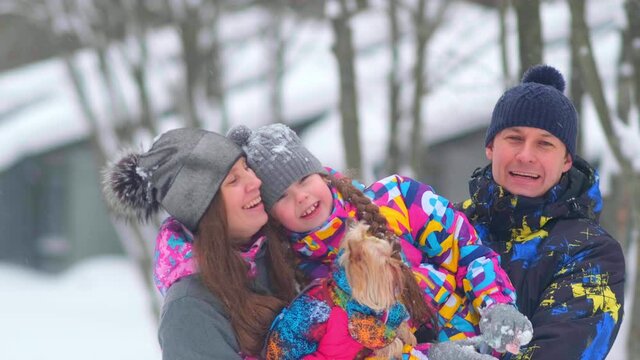happy family with little daughter and small dog hugging and smile against houses in forest on cold winter day slow motion