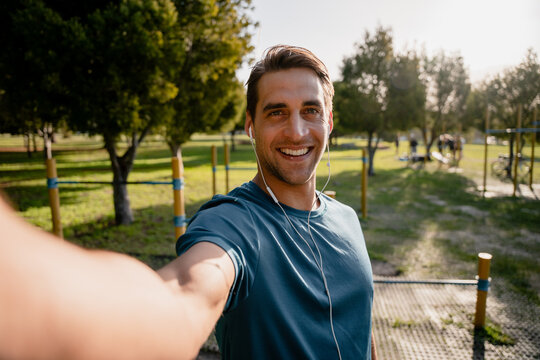 Smiling young male athlete takes photo of self before exercising outdoors in the sunny park listening to music in ear phones