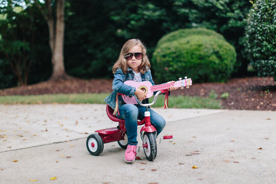 Cute young girl sitting on a tricycle while rocking out with her toy guitar