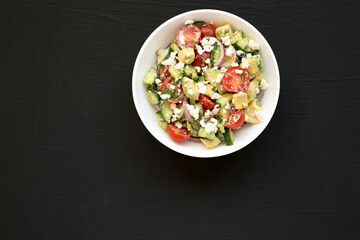 Delicious Avocado Tomato and Cucumber Salad in a white bowl on a black surface, top view. Flat lay, overhead. Copy space.