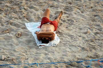 A man is lying on the beach while he holds his hands in front of his face to protect himself from the sun. A corona cordon on Villajoyosa beach in Spain.