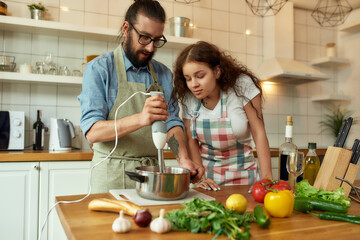 Italian man, chef cook using hand blender while preparing a meal. Young woman, girlfriend in apron...