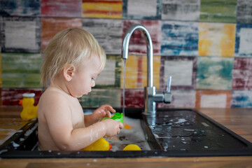 Cute smiling baby taking bath in kitchen sink. Child playing with foam and soap bubbles in sunny kitchen with rubber ducks