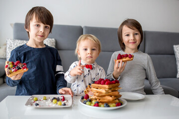 Sweet toddler birthday boy and his brothers, eating belgian waffle with raspberries, blueberries, cocnut and chocolate for breakfast