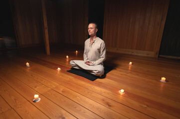 A portrait of an attractive Caucasian man sits in cotton robes in a lotus pose in a dark wooden...
