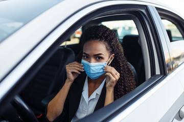 Young business woman behind the steering wheel of a taxi adjusts the medical mask. Business trips during pandemic, new normal and coronavirus travel safety concept.