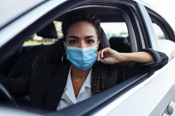Young business woman fixing and adjusting her medical mask while sitiing in the car behind the steering wheel. Business trips during pandemic, new normal and coronavirus travel safety concept.