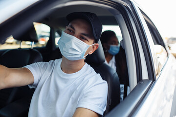 Young taxi driver works during covid-19 quarantine wearing medical mask for safety and giving a ride to a passanger. Business trips during pandemic, new normal and coronavirus travel safety concept.