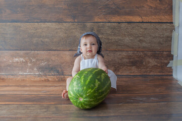 Fototapeta premium One year old girl is sitting with watermelon. She is dressed in a chef's suit. White apron. On wooden background. Summer time. Birthday photoshoot. 